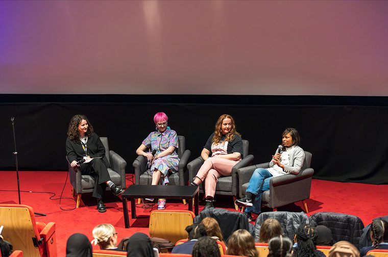 four people are seated, talking with microphones infront of a crowd.