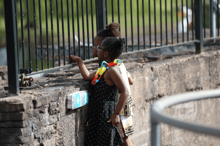 Two young girls looking through a thin metal barrier. They are stood in an outdoor theatre and there is a park on the other side of the barrier. One of the young girls has a paper garland around her neck.