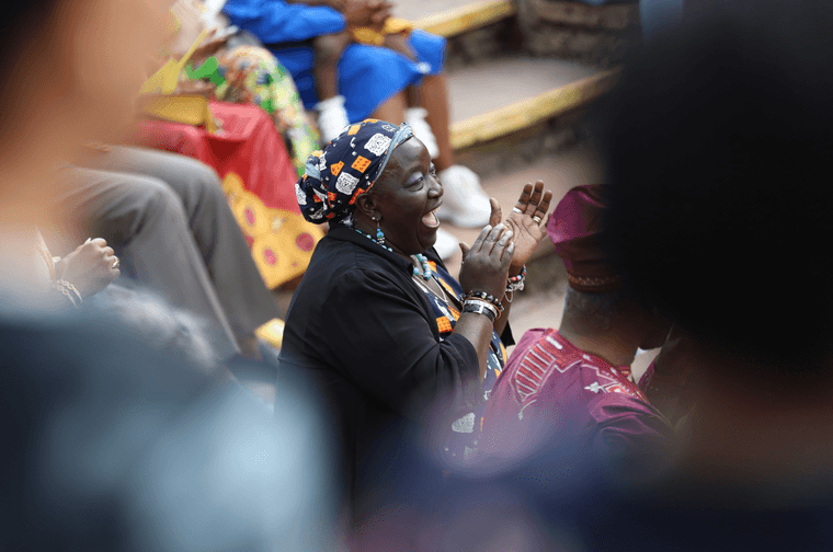 A close up of a woman in a crowd. She is sat in an outdoor theatre, smiling and clapping.