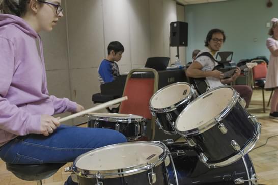 Young girl plays the drums in a music studio, other children play instruments in the background