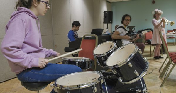 Young girl plays the drums in a music studio, other children play instruments in the background