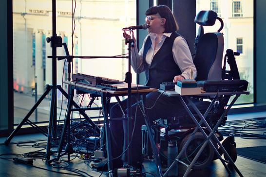 Woman sings into a microphone with keyboard and music technology. The woman is in a wheelchair, with cropped brown hair and glasses in a white shirt and black waistcoat.