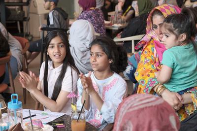 Young girls clapping in busy café and mum and smaller child on her hip wearing bright colours smiling