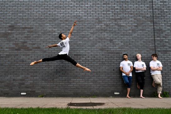 A male dancer jumps high in the air with his legs split and arms stretched out, as three more men wearing the same white t shirt and the dancer stand and watch leaning against a brick wall.