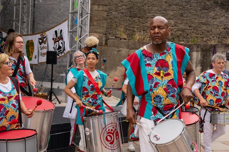 Group of people wearing teal and red tabards and playing drums which are on a rope around their necks. There are in an outdoor theatre. The drums are silver and have a red top.