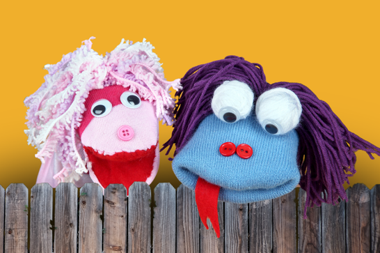 Two sock puppets with googly eyes - one pink and one blue - look down over the top of a wooden fence in front of a yellow background.