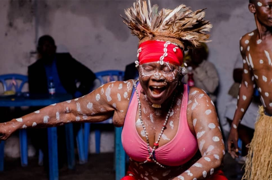 A dancer has a happy expression with their eyes closed and move open and their hair is moving above their head - they wear a read headband and has spots of white paint over their arms and face.