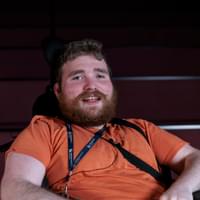 A smiling man in his twenties with a beard smiles at the camera. He is sitting in a wheelchair, wearing a lanyard and an orange T-shirt.