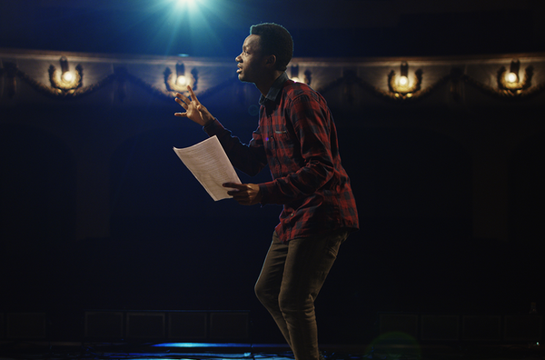 Image shows a young black man with short hair, wearing a red and black plaid shirt and grey jeans. He is in a slightly crouched position on stage against a black, dimly lit theatre background.
