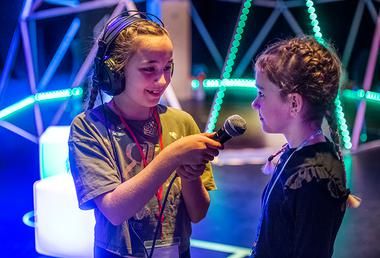 A young girl wearing headphones holds up a microphone for another young girl