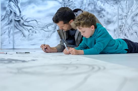 A man and a young boy lay on their stomachs side by side while drawing