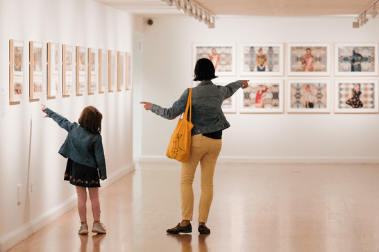 A woman and a little girl point at photography on bright white gallery walls