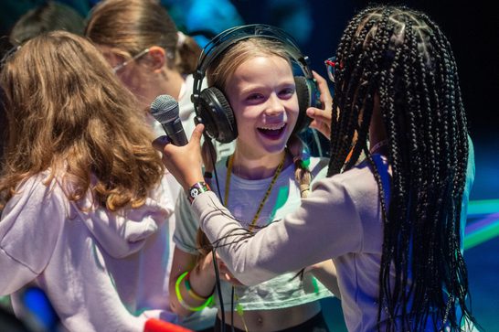 A young girl with braids puts headphones on another girl with a wide smile