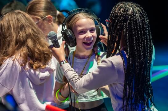 A young girl with braids puts headphones on another girl with a wide smile