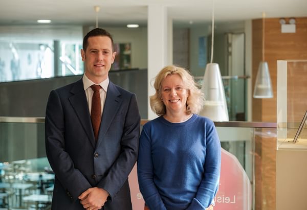 Man in a suit and woman in a blue jumper stand next to each other in an arts centre, smiling.