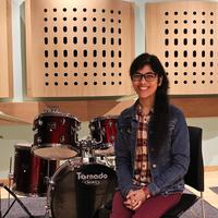 A smiling woman with long dark hair and glasses sitting by a drum kit