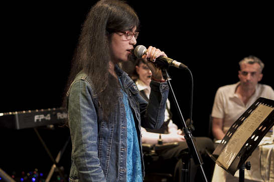 A woman with dark hair and glasses singing into a microphone