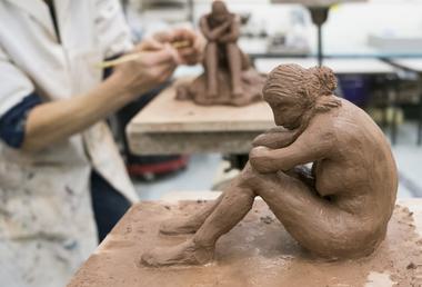 Clay sculpture of a woman sat with arms crossed leaning on her knees with an artist's hands in the background