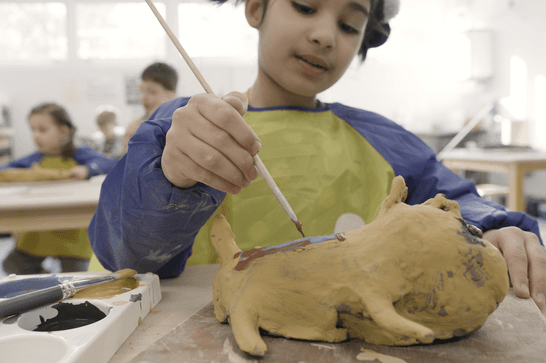 A young girl in a yellow smock painting a clay sculpture of an animal