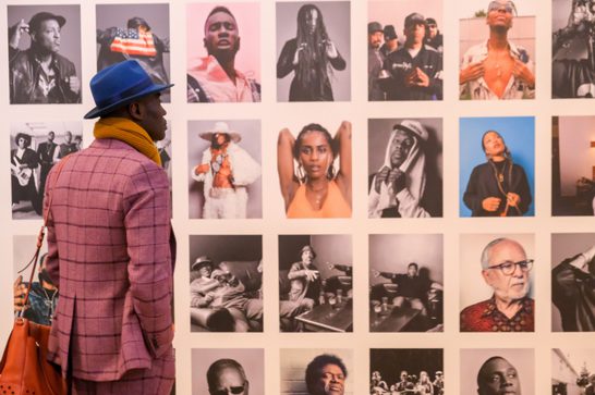 A man in checked pink suit and blue hat looks at portraits on a gallery wall
