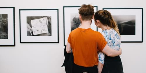 A young man with his arm around a young woman, looking at photography on a gallery wall