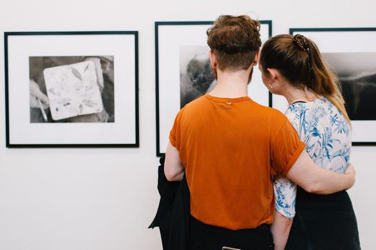 A young man with his arm around a young woman, looking at photography on a gallery wall