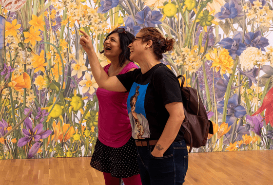 Two women stand in front of a floral mural smiling and pointing at a piece of art