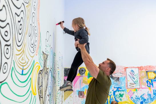 a man is holding a small child up above his head, who is using a big pen to drawing funky patterns on a wall.