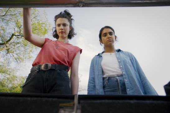 Two young women look into the trunk of a car, one of them holding up the lid. Green foliage is in the background.