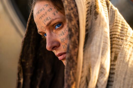 A woman in a hood (Rebecca Ferguson) with piercing blue eyes looks out to the left of the picture. Strange symbols and Cyrillic writing cover her face.