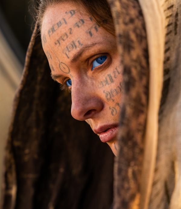 A woman in a hood (Rebecca Ferguson) with piercing blue eyes looks out to the left of the picture. Strange symbols and Cyrillic writing cover her face.