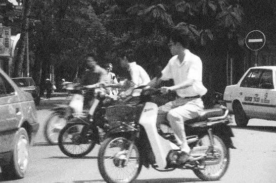 Three Vietnamese men ride motorbikes, seen in black and white.