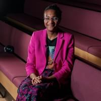 A younger Black woman wearing glasses smiles at the camera. She is wearing a pink blazer and a flowery skirt and is sat on a purple cushioned pew.