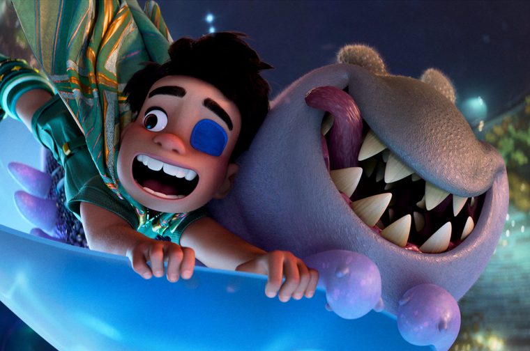 A small boy with a blue eye patch over his left eye holds onto the edge of a space sled. Next to him is a grey worm like alien with sharp fangs. They are speeding through space, both of them smiling.