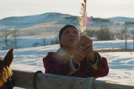 An elderly Native American man holds up a feather. He is stood by a fence with a horse tied to it and is surrounded by a wintry landscape.