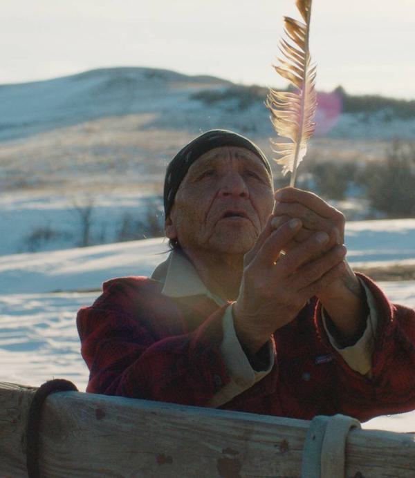 An elderly Native American man holds up a feather. He is stood by a fence with a horse tied to it and is surrounded by a wintry landscape.