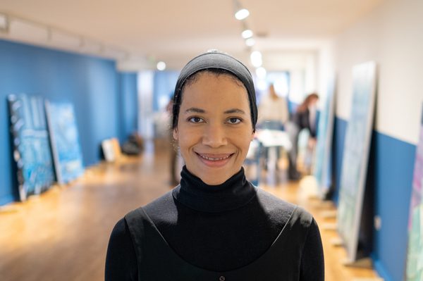 Image shows a young black woman wearing a black headscarf (tied up) and a black high-neck top. She is smiling into the camera and stands in a long, corridor-like gallery space, which has deep blue and white walls and pale wood flooring.