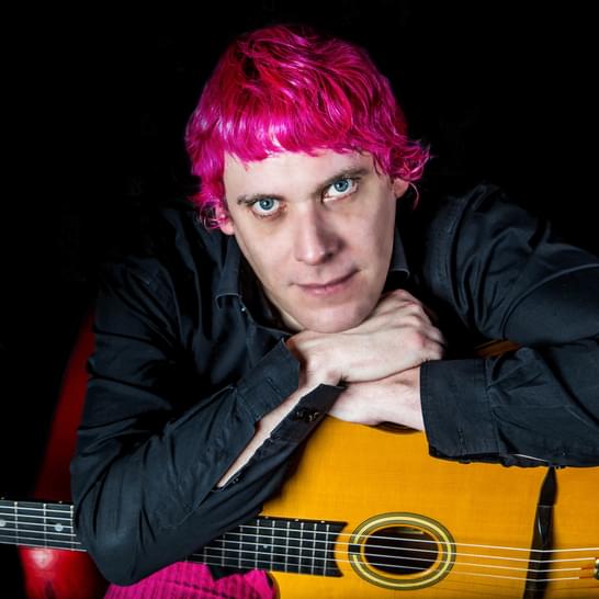 Gary is leaning onto his crossed arms, which are placed over a guitar. Gary has short pink hair.