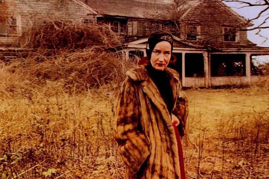 An older woman dressed in a fur coat and wearing a shawl stands in front of a dilapidated house surrounded by weeds.