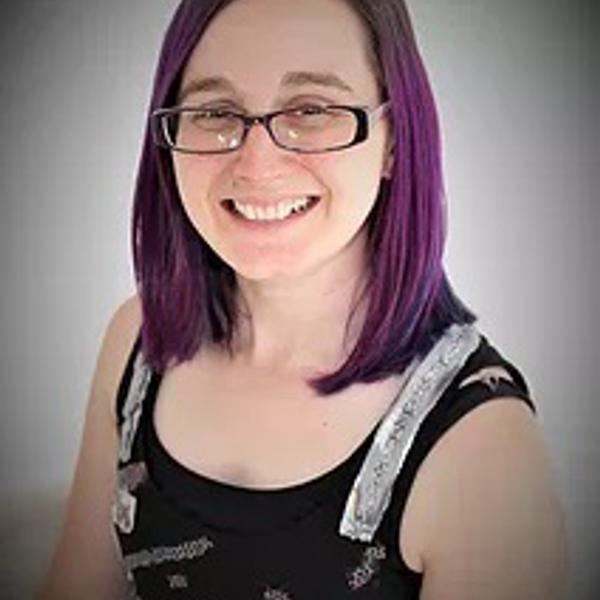 Helen is sat against a white background and has purple hair. Helen is wearing a black tank top and glasses.