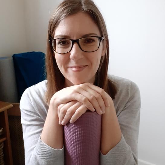 Helen has her hands placed over a purple yoga mat. Helen has long brown hair and is wearing glasses.