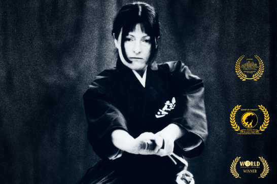 Black and white: a woman wearing martial arts robes and with her hands on the hilt of a sheathed sword.