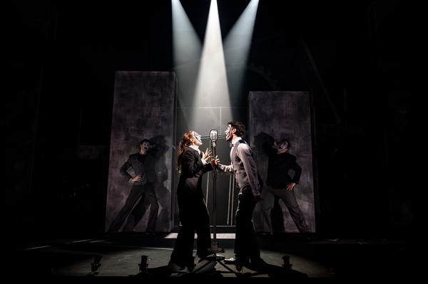 Two actors on stage singing into a microphone facing each other. Dramatic lighting points onto the microphone. In the background dancers are posed.