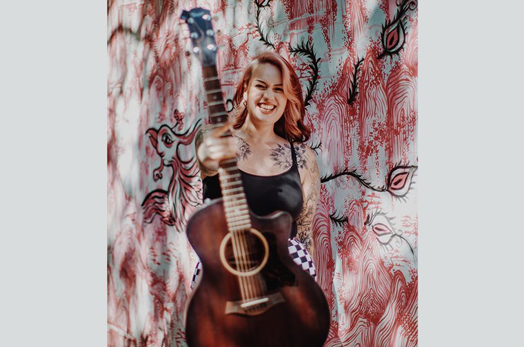 Image of Lily Kerbey holding guitar towards the camera and smiling with pink graffiti behind her on the wall
