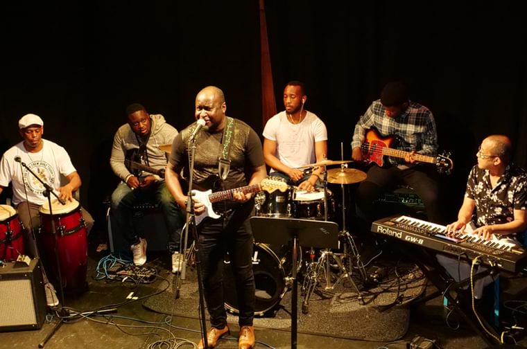 6 men playing instruments on a dark stage. The man in the middle is playing an electric guitar and singing into a microphone.