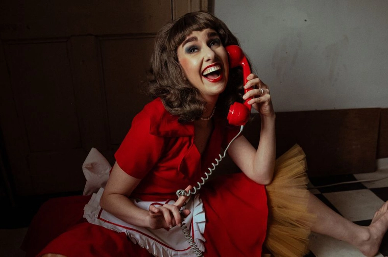 Woman with brunette hair styled in 50’s bouffante wearing a red dress laughing while on a corded red telephone