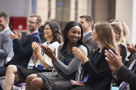 A group of people sitting in a row of chairs wearing smart work clothes, clapping and smiling