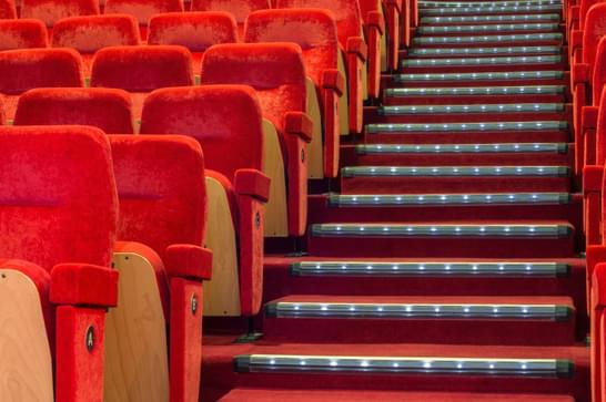 To the left, rows of red cinema seats are located with the bottom of the seats folded up. To the right, there are steps going up with the same colour red fabric covering them.