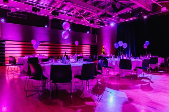 A room with balloons, tables and chairs is lit up with pink lighting