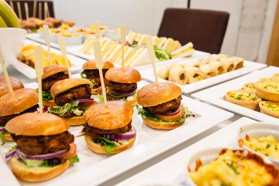 A collection of small plates, with mini hamburgers to the left, mini quiche to the right and cut sandwiches in the background.